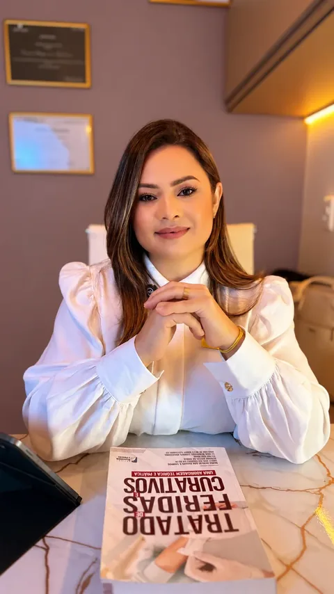 Dra. Daiana Lima no consultório, com o livro Tratado de Feridas e Curativos sobre a mesa e certificados ao fundo